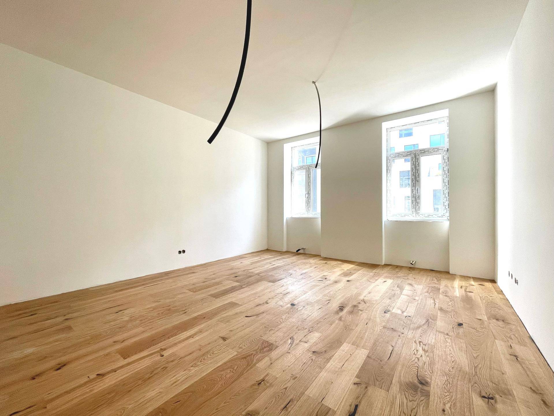 Heller, frisch renovierter Raum mit neu verlegtem Holzparkettboden und zwei Fenstern.