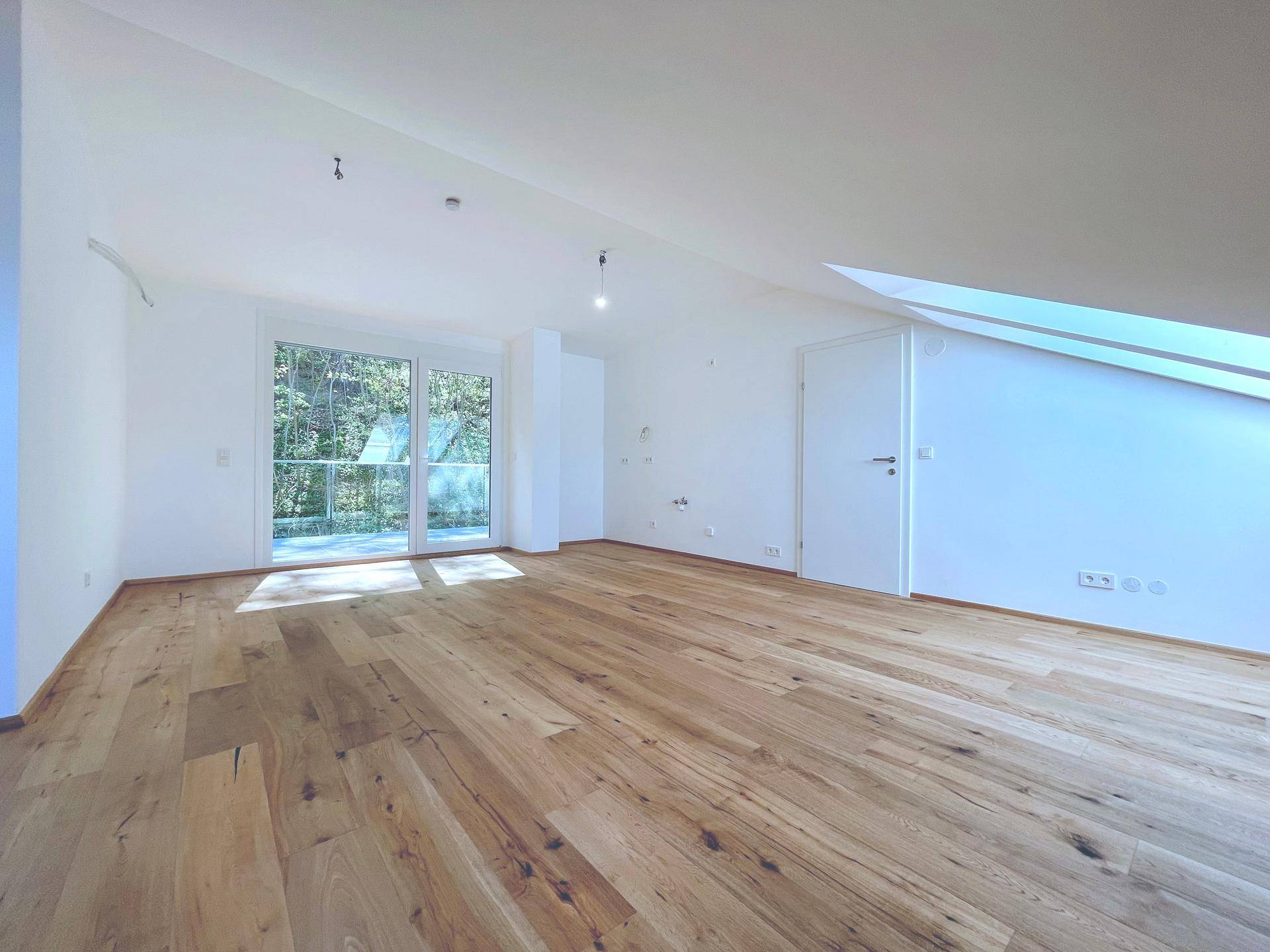 Lichtdurchflutetes Wohnzimmer im Dachgeschoss mit Holzboden und großen Fenstern, bietet einen grünen Ausblick.