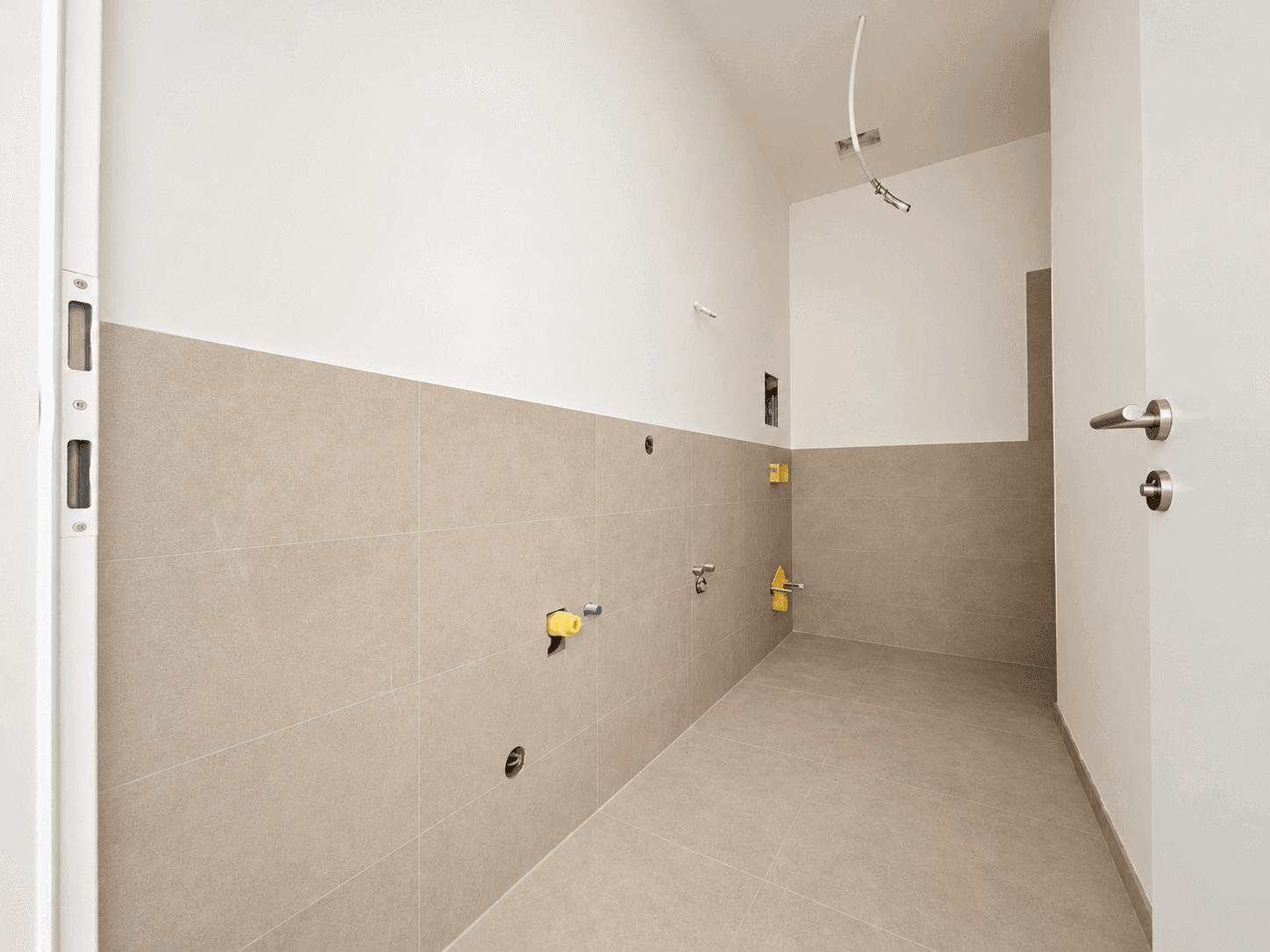 Unfertiges Badezimmer mit beigefarbenen Fliesen und sichtbaren Wasseranschlüssen, bereit für die Installation.