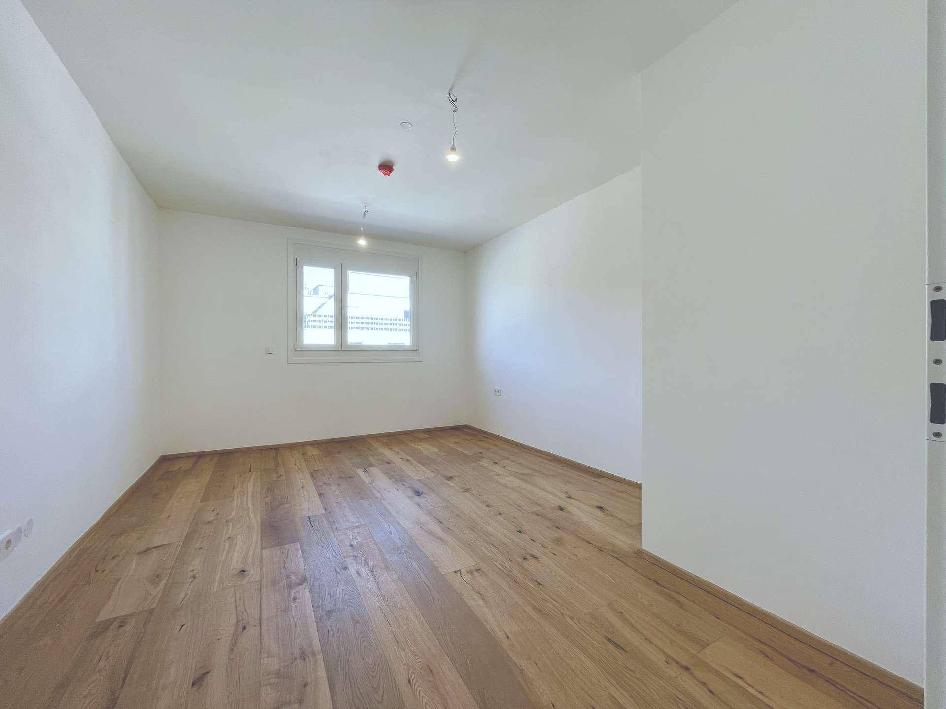 Helles, leeres Schlafzimmer mit Holzboden und einem Fenster, bereit zur Einrichtung.