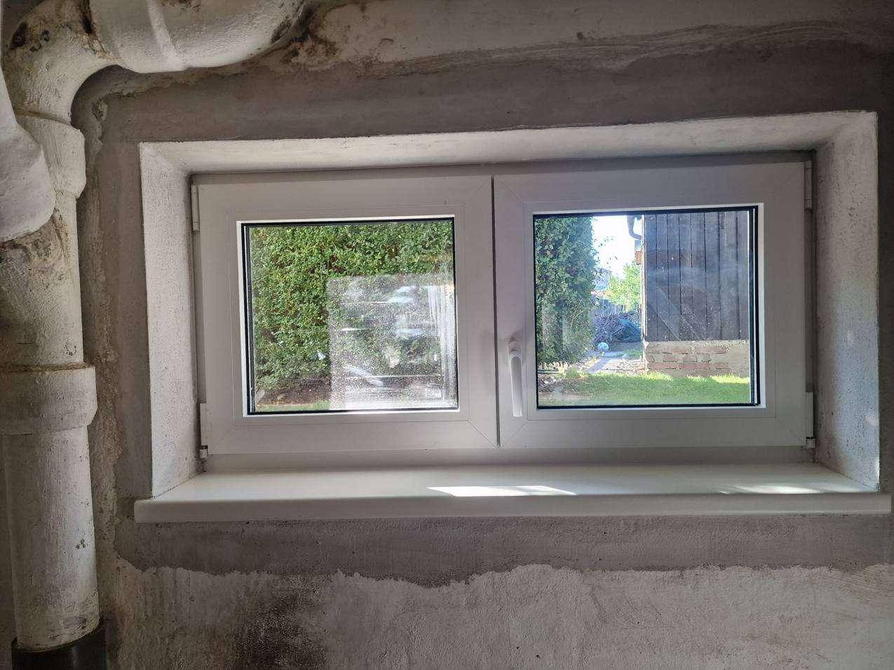 Kleines Fenster in einem unverputzten Raum mit Blick ins Grüne und sichtbaren Rohren.