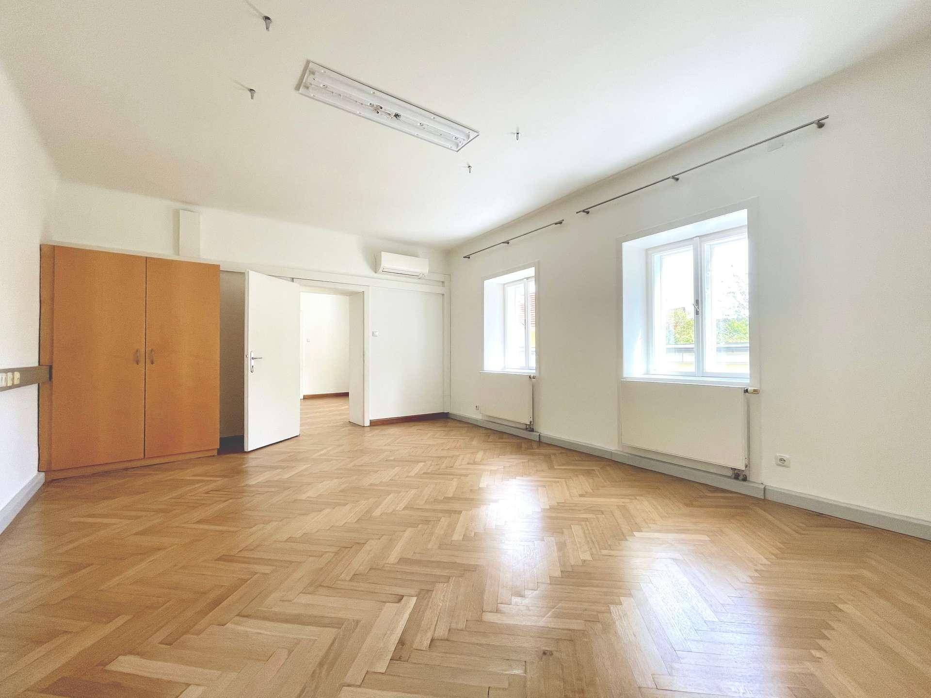 Heller Raum mit Fischgrätparkett, zwei Fenstern, Klimaanlage und einem Holzschrank.