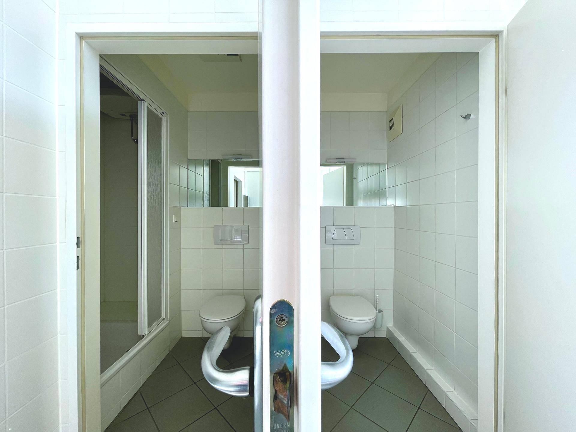 Zwei separate Toilettenbereiche mit weißen Fliesen, Dusche und modernen Wand-WCs.