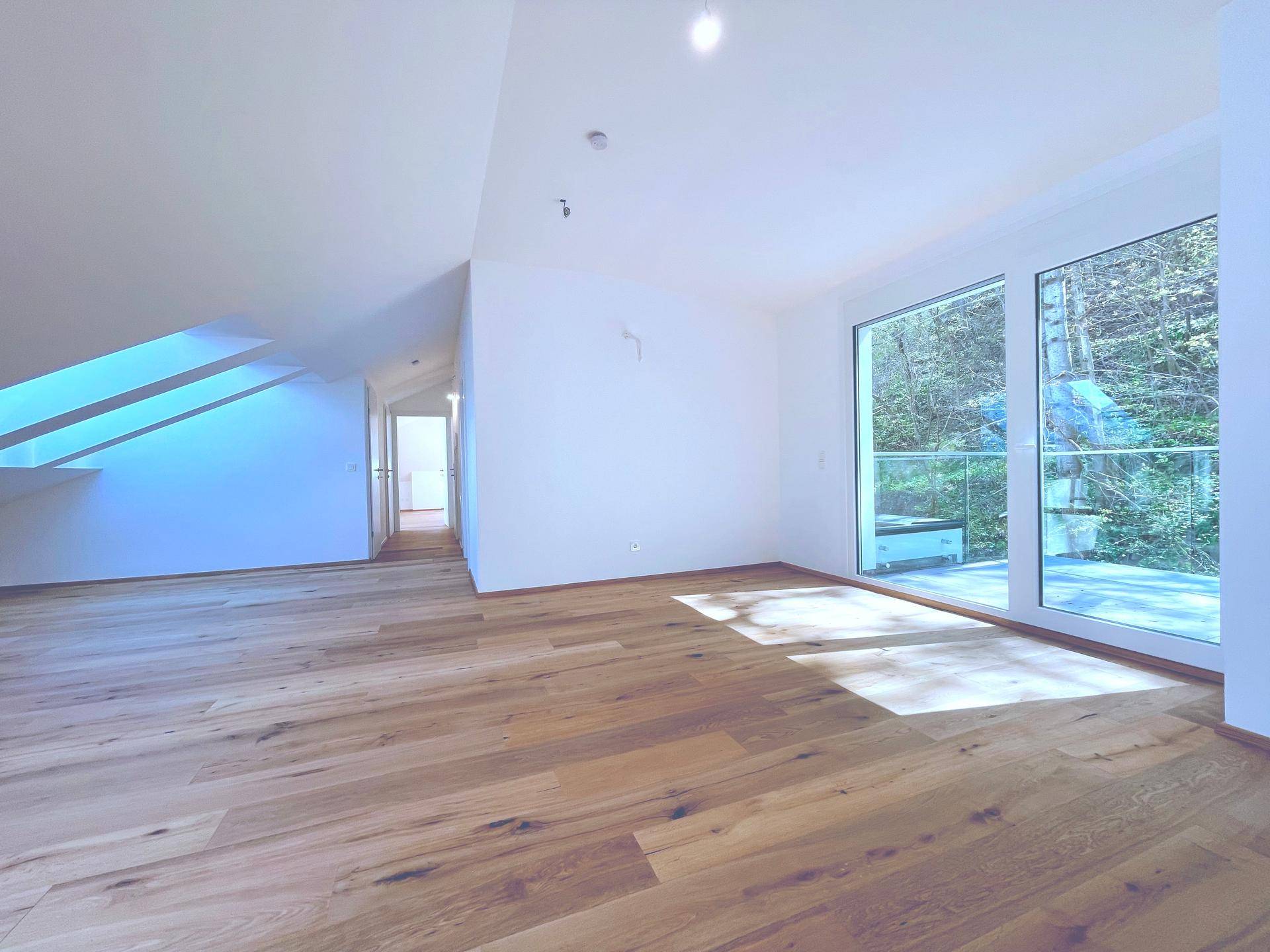 Geräumiges Wohnzimmer im Dachgeschoss mit Holzboden und großen Fenstern, bietet einen grünen Ausblick.