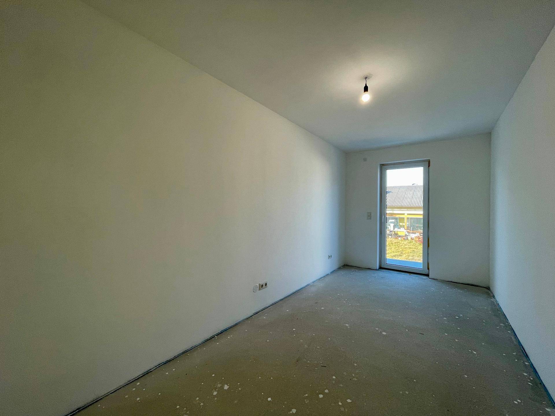 Unmöbliertes Zimmer im Rohbau mit einer Fenstertür und Betonboden.
