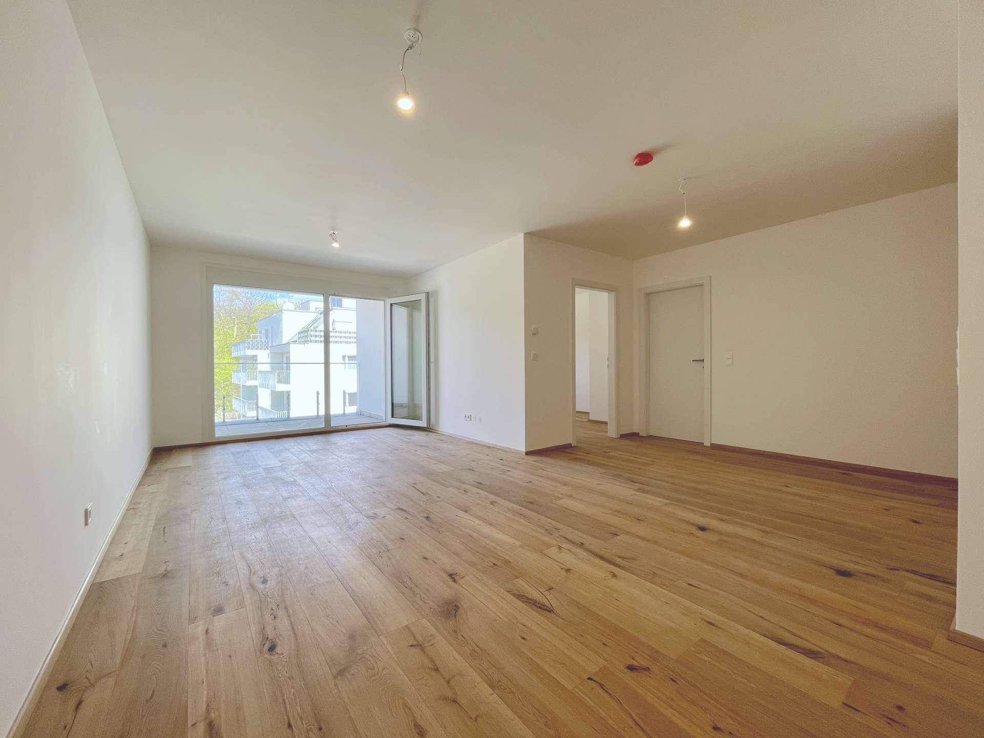 Geräumiges, leeres Wohnzimmer mit Holzboden und großen Fenstern, die viel Licht hereinlassen.