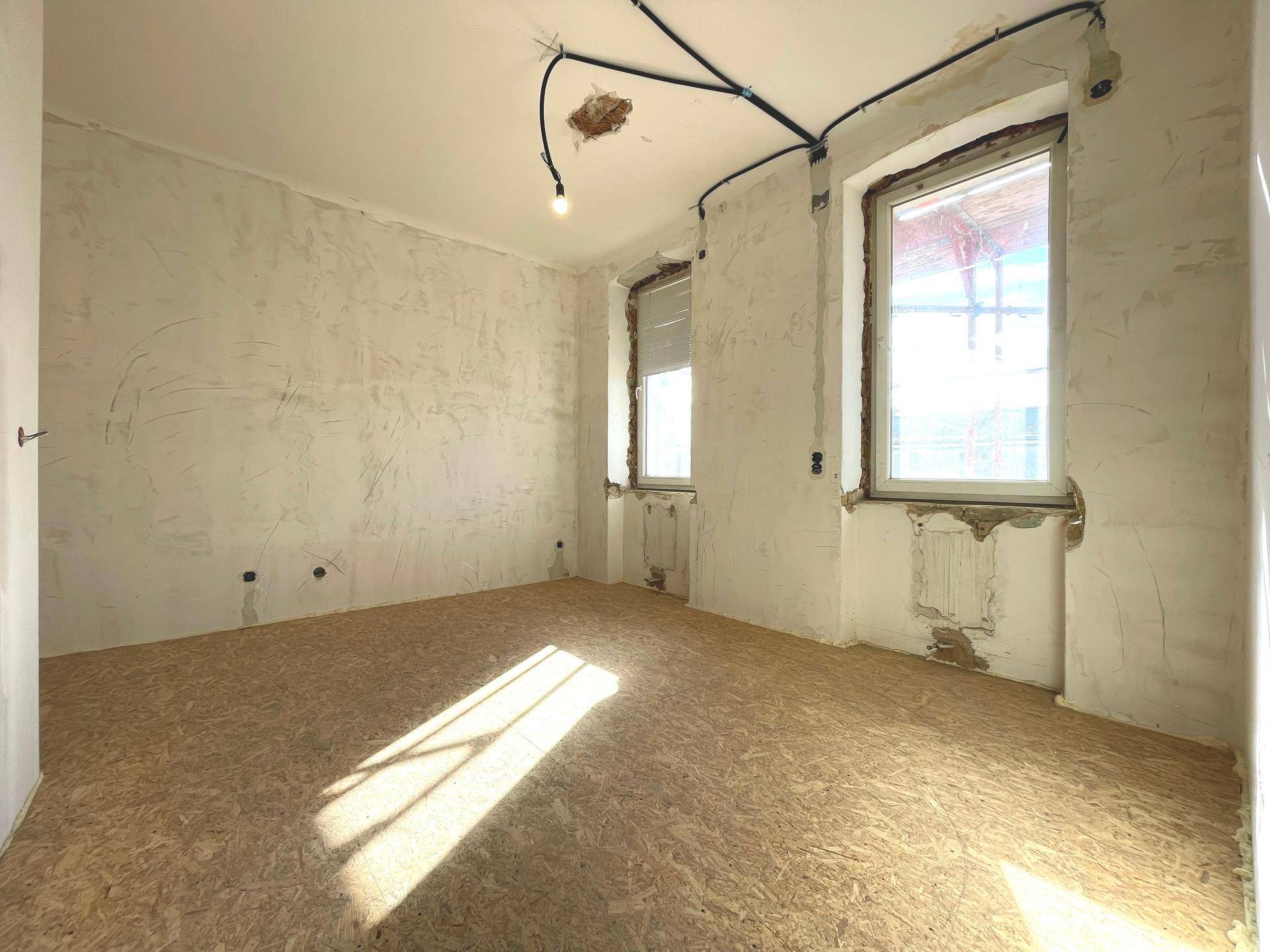 Raum im Rohbau mit sichtbaren Leitungen und Fenstern, bereit für die Fertigstellung.
