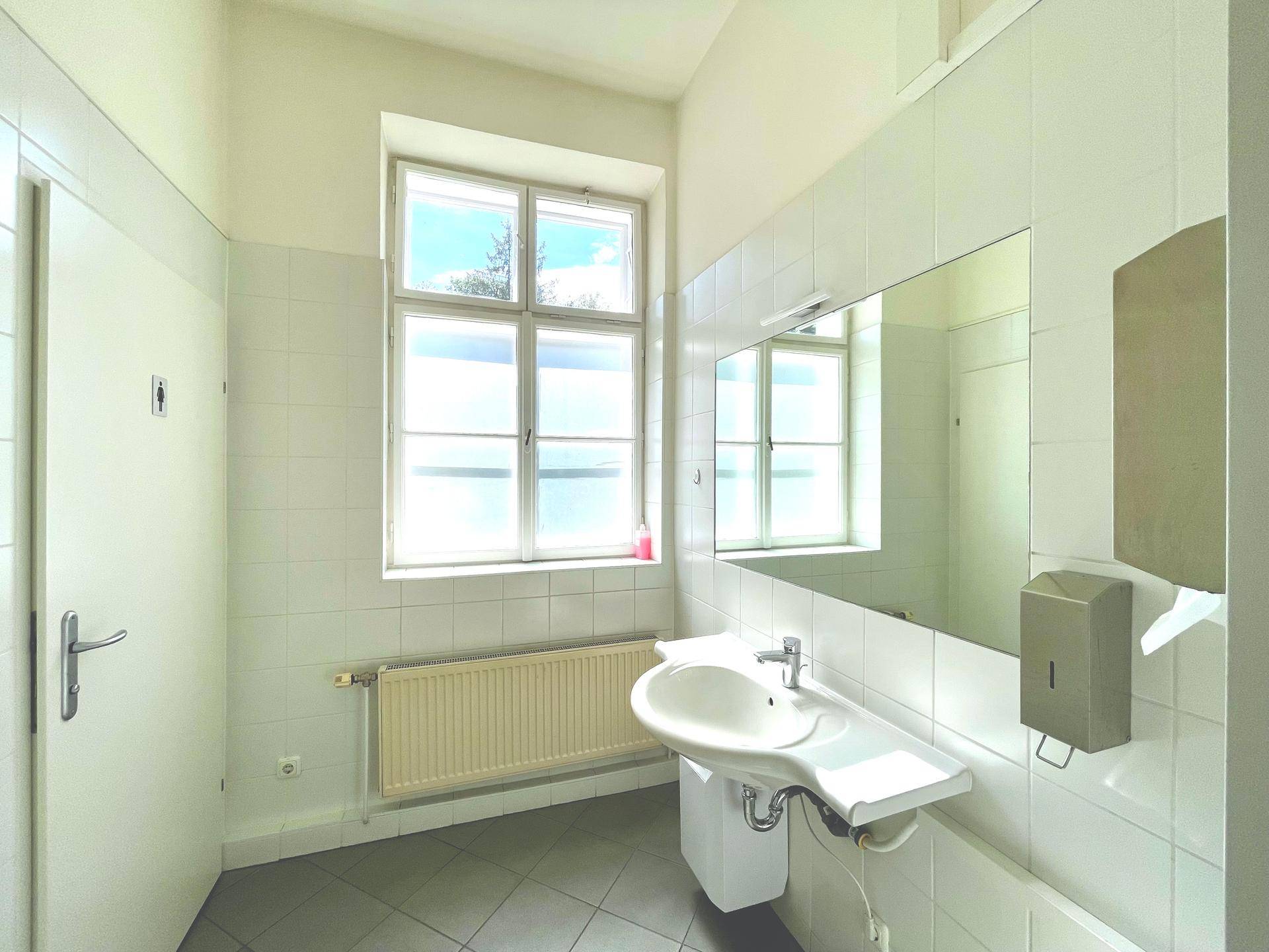 Helles Badezimmer mit weiß gefliesten Wänden, Fenster und einem Waschbecken mit Spiegel.