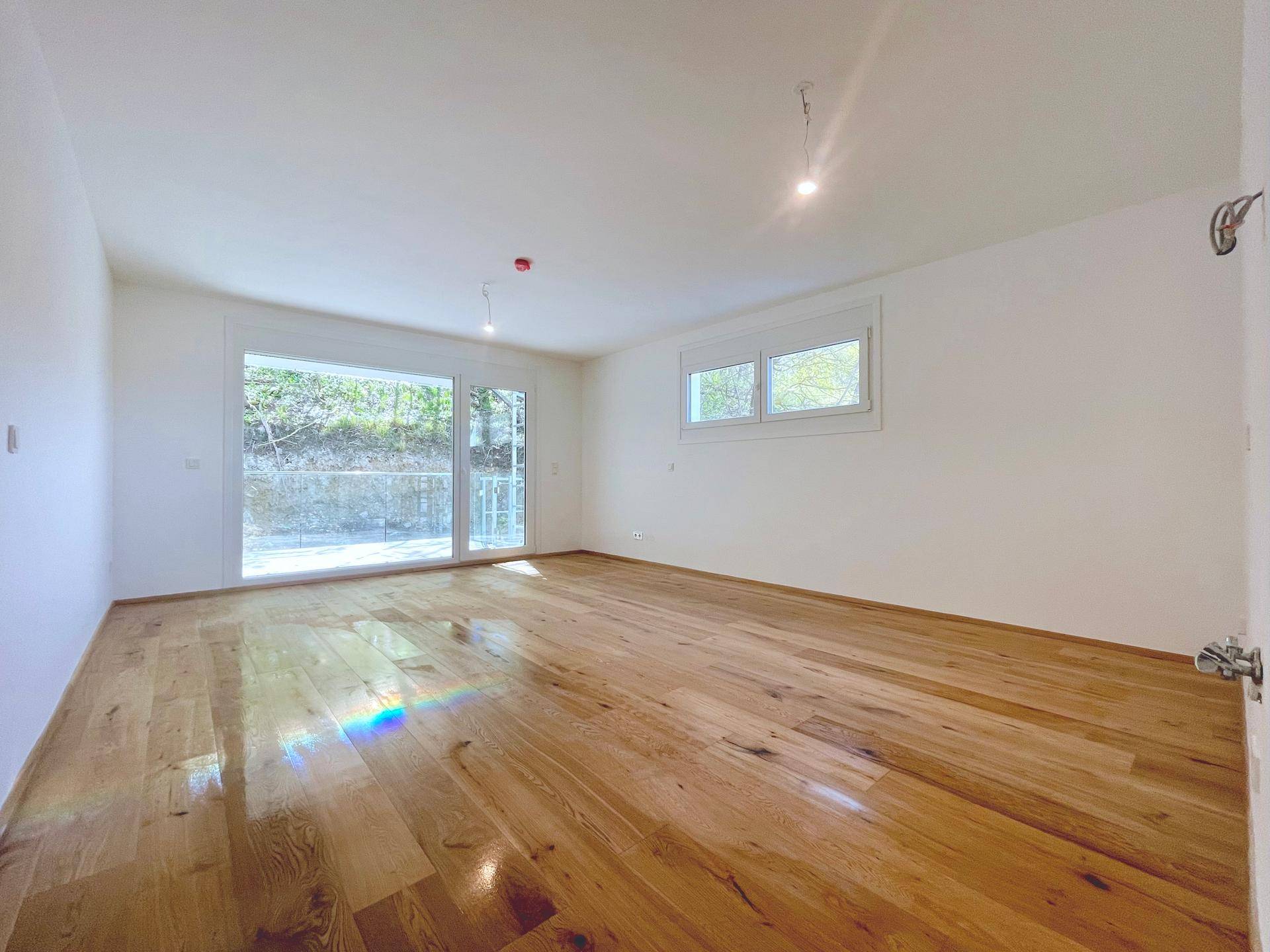 Geräumiges Wohnzimmer mit hellem Holzboden und großen Fenstern, bietet einen grünen Ausblick.