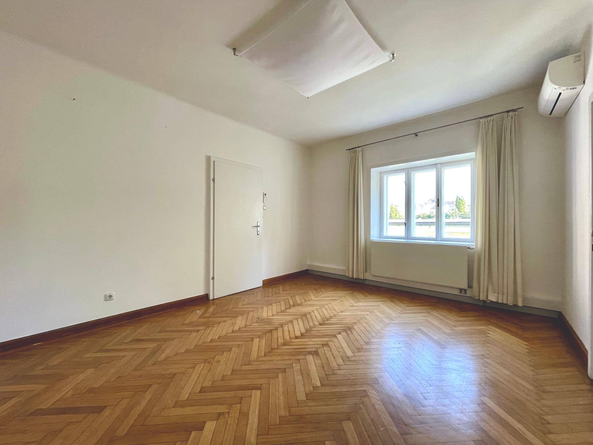 Heller Raum mit Fischgrätparkett, einem großen Fenster und einer Klimaanlage an der Wand.