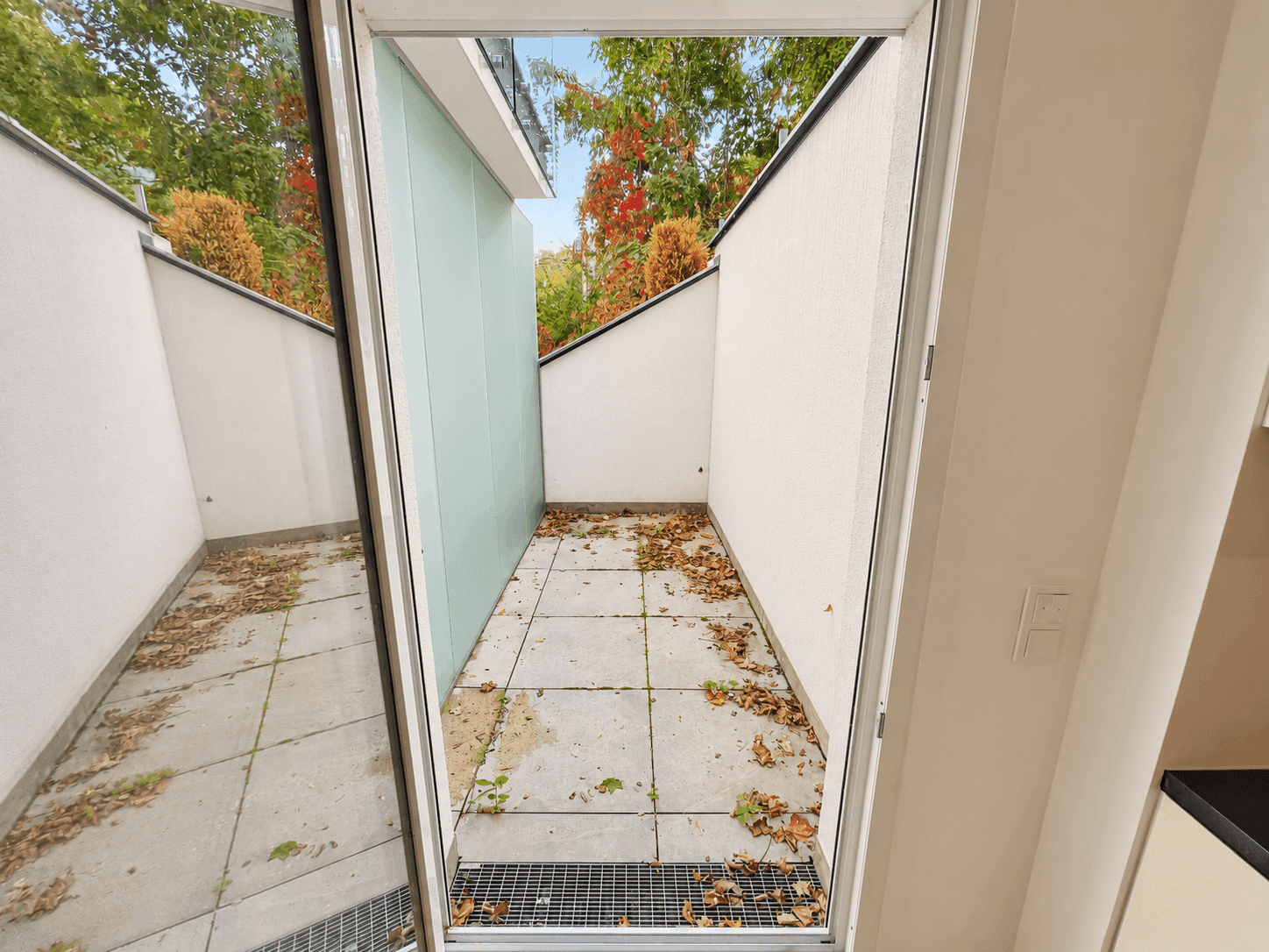 Kleine, geflieste Terrasse mit seitlicher Glasabtrennung und herbstlichem Laub auf dem Boden.