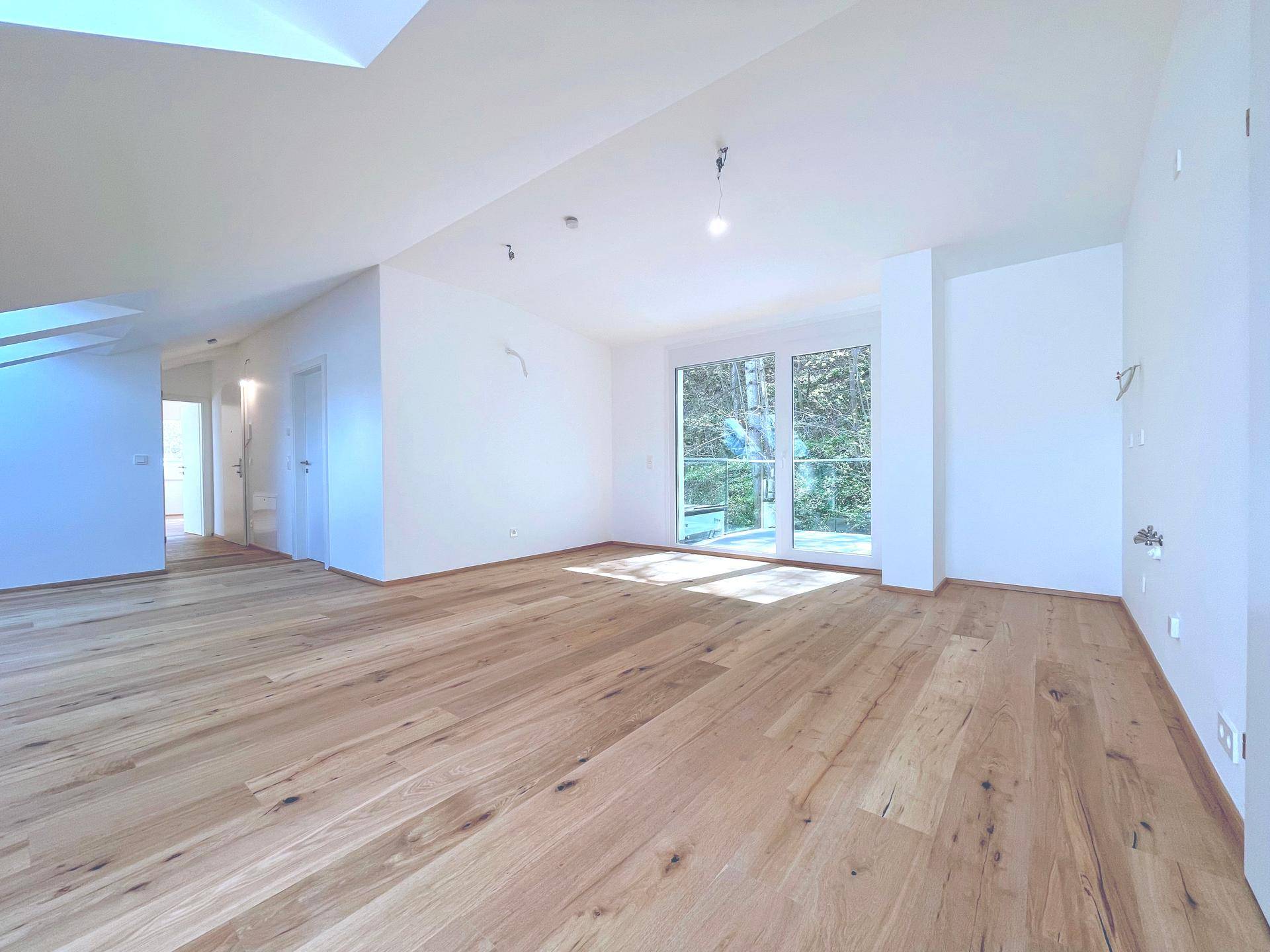 Helles Wohnzimmer im Dachgeschoss mit Holzboden und großen Fenstern, bietet einen grünen Ausblick.