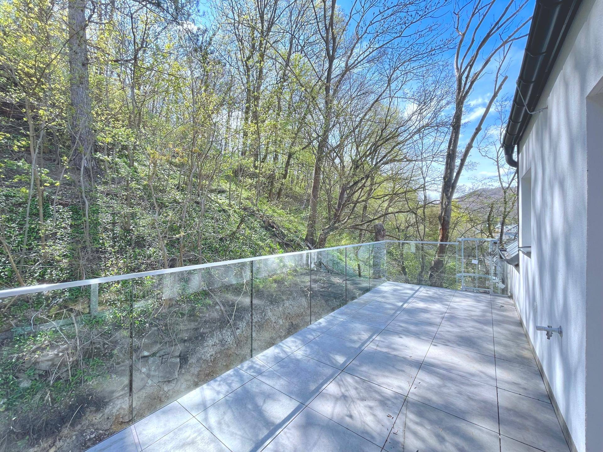 Schmaler Balkon mit Fliesenboden und Glasgeländer, bietet einen grünen Blick auf die umliegenden Bäume.