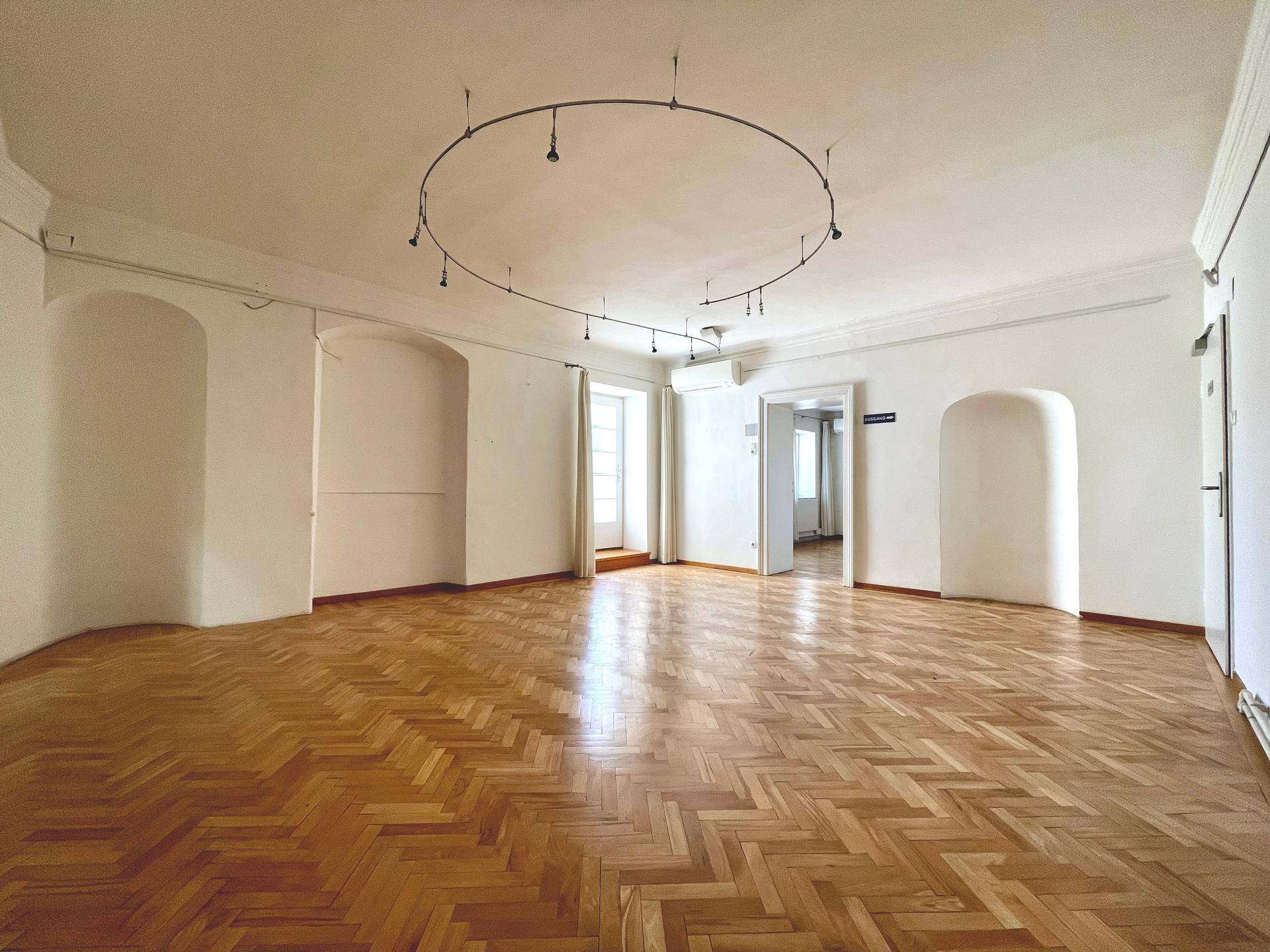 Großer Raum mit Fischgrätparkett, hohen Decken und einem Fenster mit Zugang zum Balkon.