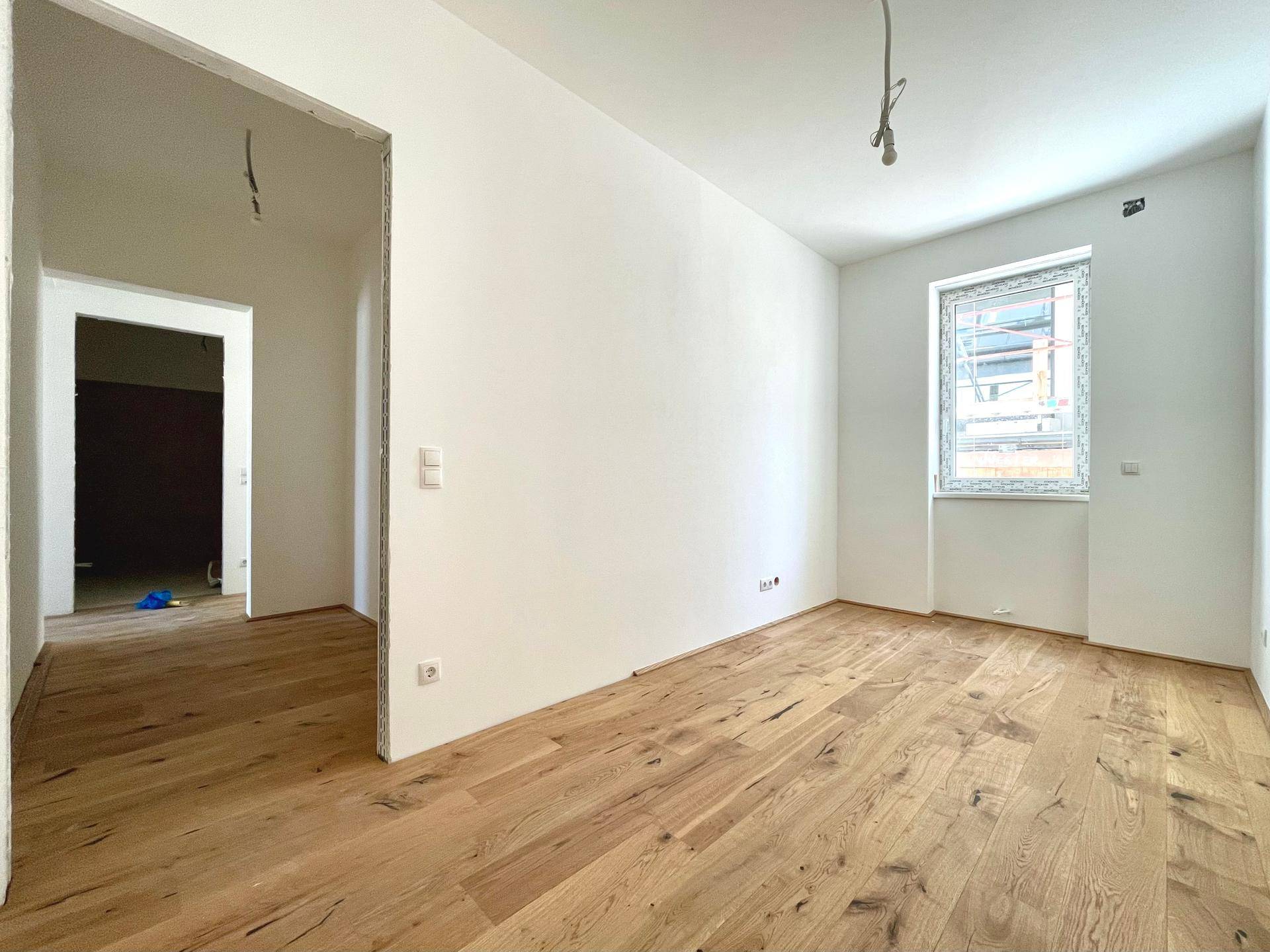 Heller Raum im Rohbauzustand mit neu verlegtem Holzdielenboden und einem Fenster.