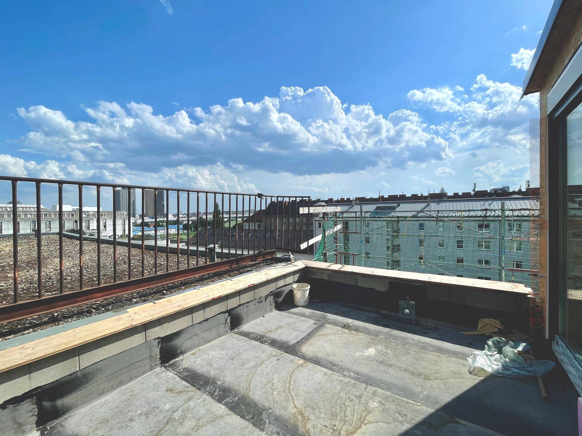 Unfertige Dachterrasse mit Metallgeländer bietet einen weiten Blick über die städtische Umgebung unter blauem Himmel.