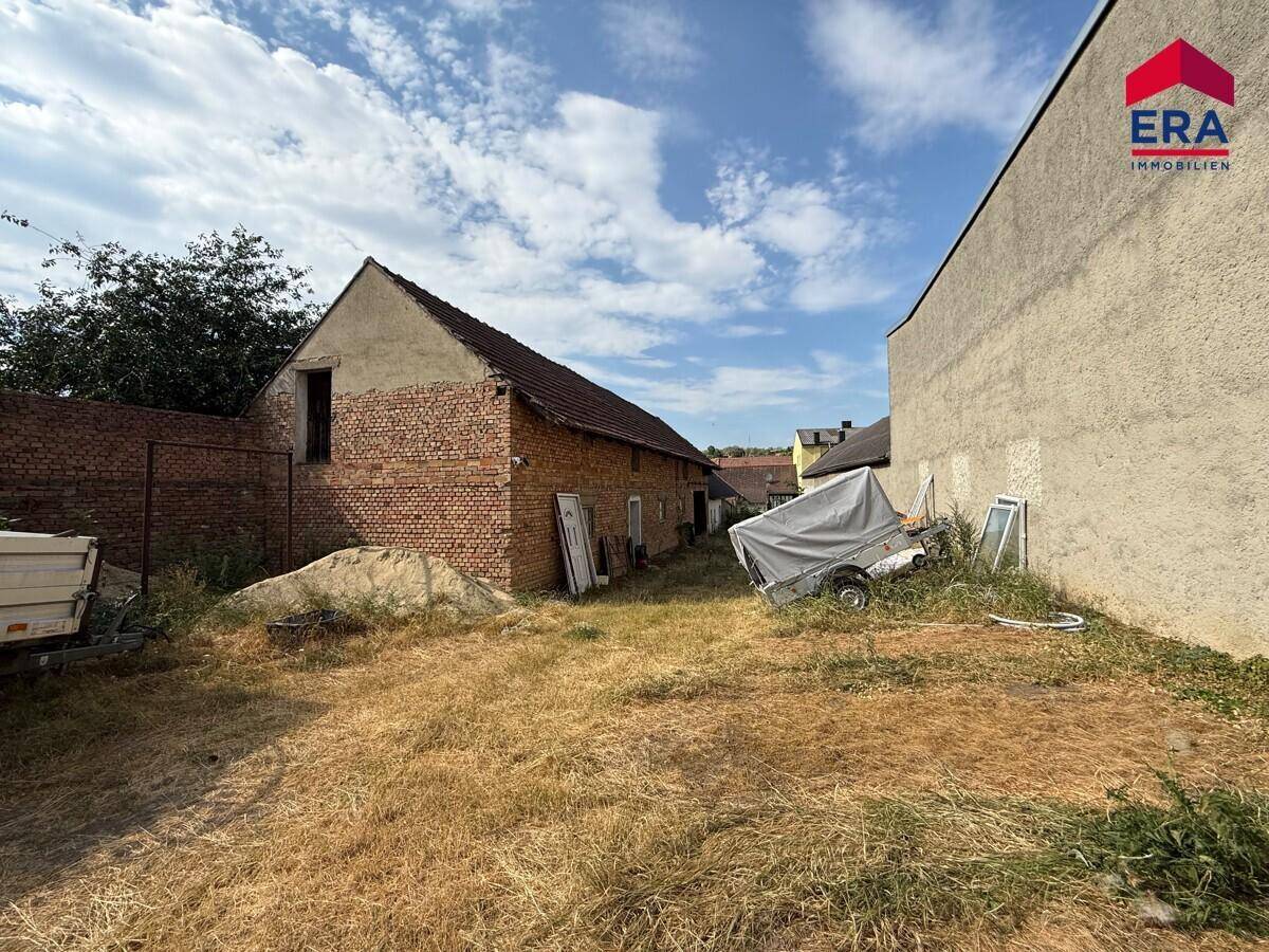 Seitenansicht der Scheune mit Ziegelwänden und einem Anhänger auf dem trockenen Gras.