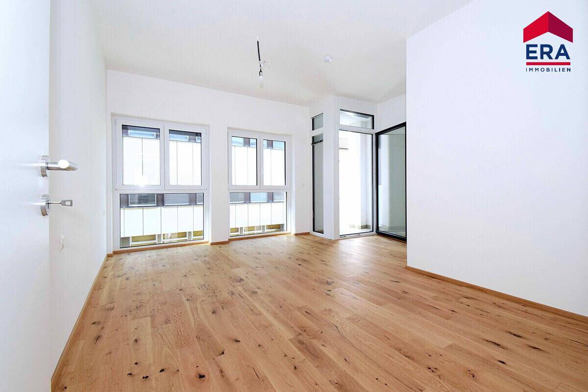 Heller Raum mit Holzboden, großen Fenstern und Zugang zu einem französischen Balkon.