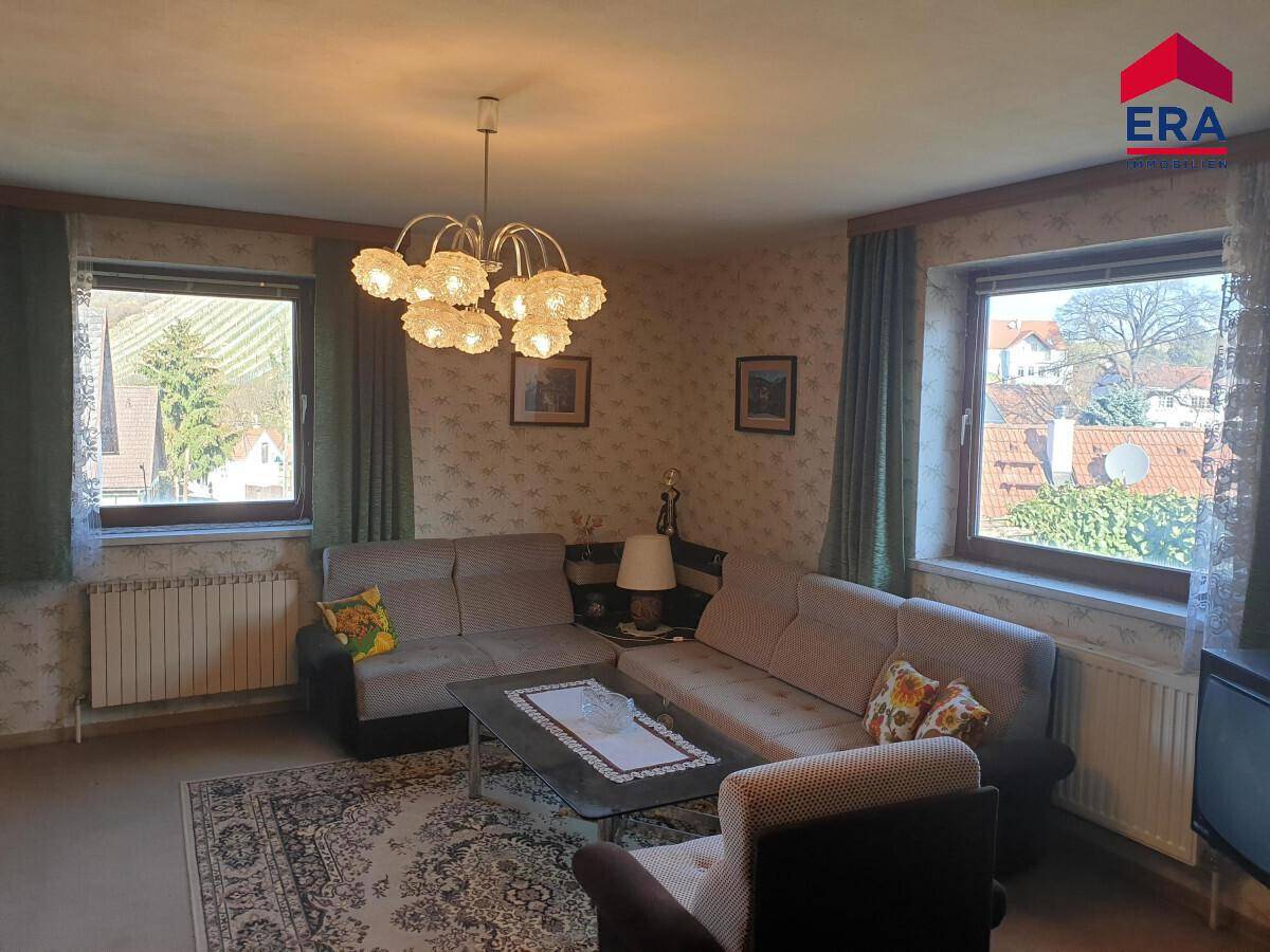 Helles Wohnzimmer mit Ecksofa, Teppichboden und zwei Fenstern mit Blick auf die Umgebung.