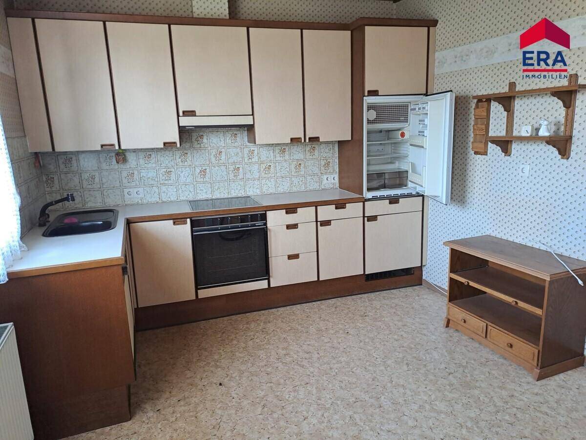 Traditionelle Küche mit hellen Holzfronten, integrierten Geräten und gemusterten Tapeten im 80er-Jahre-Stil.