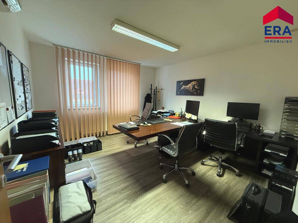Geräumiges Büro mit mehreren Schreibtischen, Bürostühlen und einem Fenster mit Jalousien.