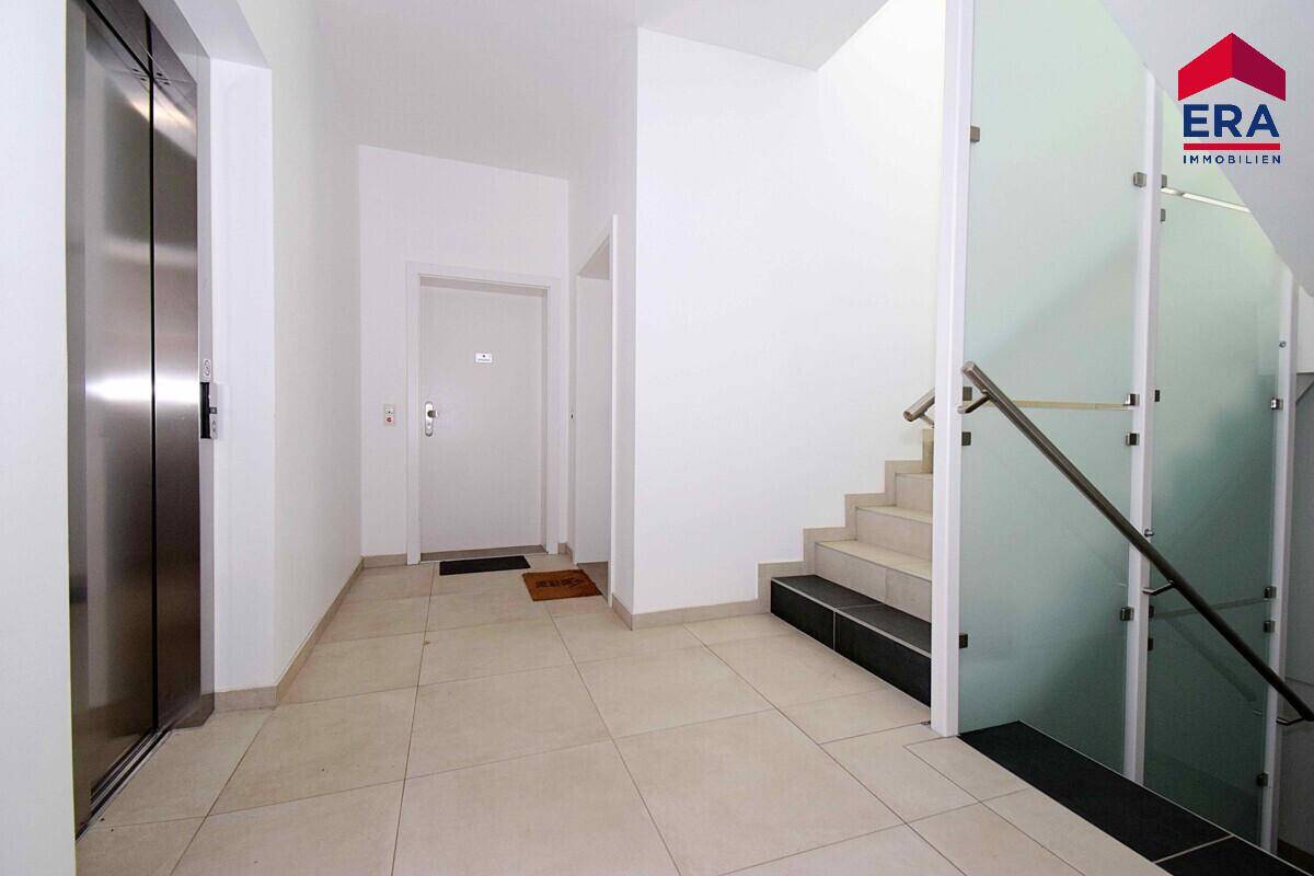 Heller Eingangsbereich mit Fliesenboden, Aufzug und Treppenhaus mit Glasgeländer.