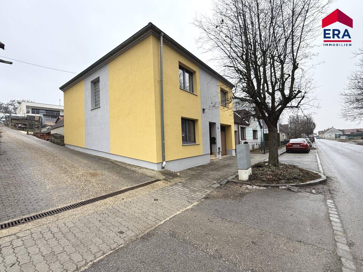 Seitenansicht des Hauses mit gelb-grauer Fassade, angrenzender Straße und einem kahlen Baum.