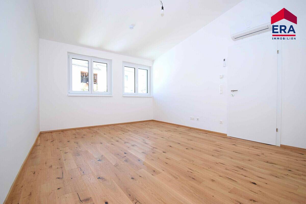 Helles Schlafzimmer mit Holzboden, zwei Fenstern und Klimaanlage.