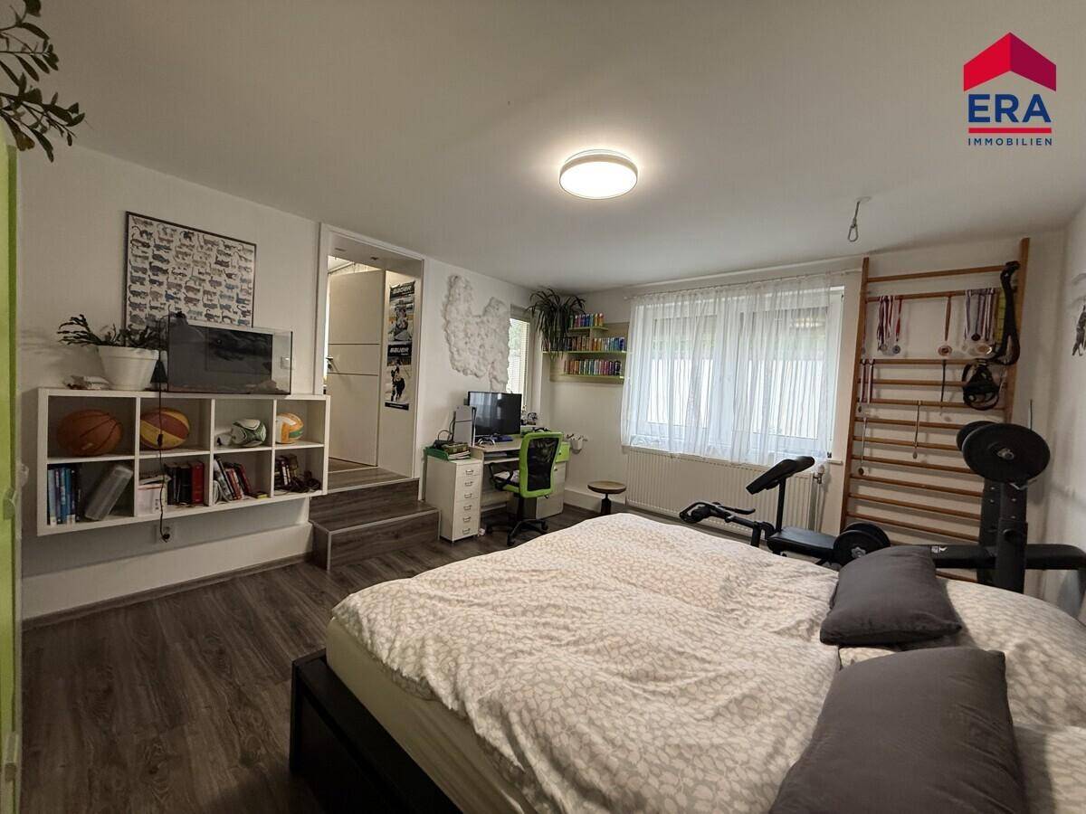 Schlafzimmer mit Bett, Schreibtischbereich und Fitnessgeräten, ausgestattet mit Laminatboden und einem großen Fenster.