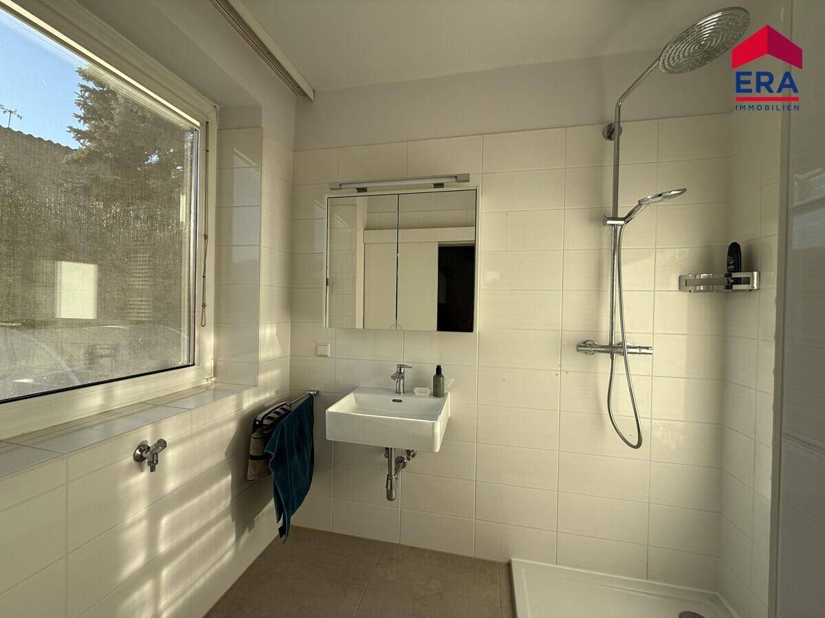 Helles Badezimmer mit Dusche, Waschbecken, Spiegelschrank und einem großen Fenster.