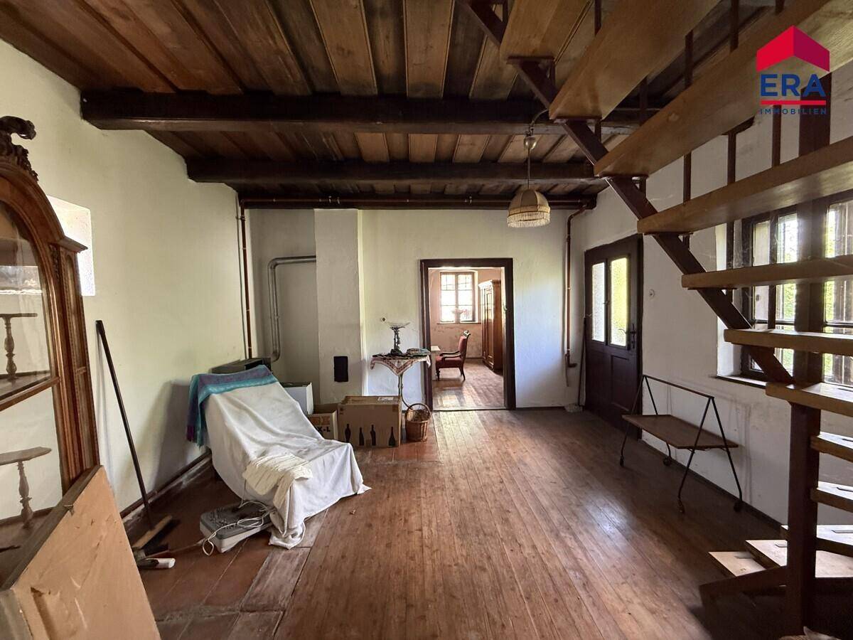 Rustikaler Innenraum mit Holzboden, sichtbaren Deckenbalken und einer Holztreppe, die in den oberen Bereich führt.