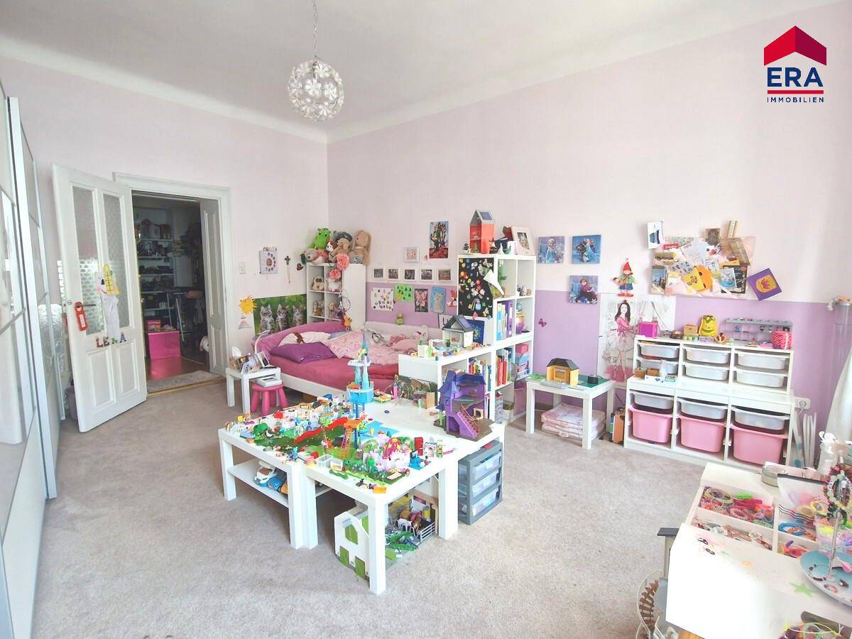 Geräumiges Kinderzimmer mit rosa Wänden, Teppichboden und vielen Spielsachen und Regalen.