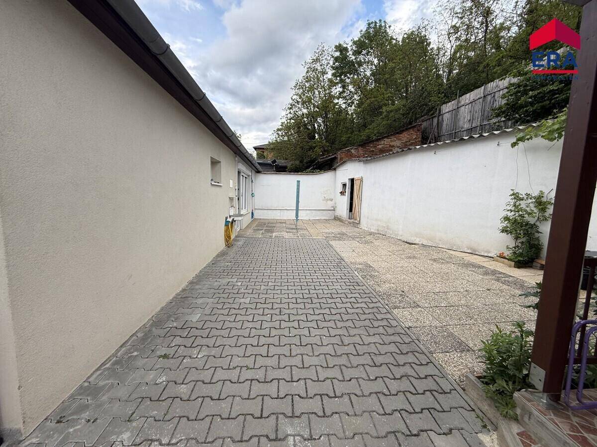 Langer gepflasterter Weg zwischen Haus und Mauer, umgeben von Bäumen im Hintergrund.