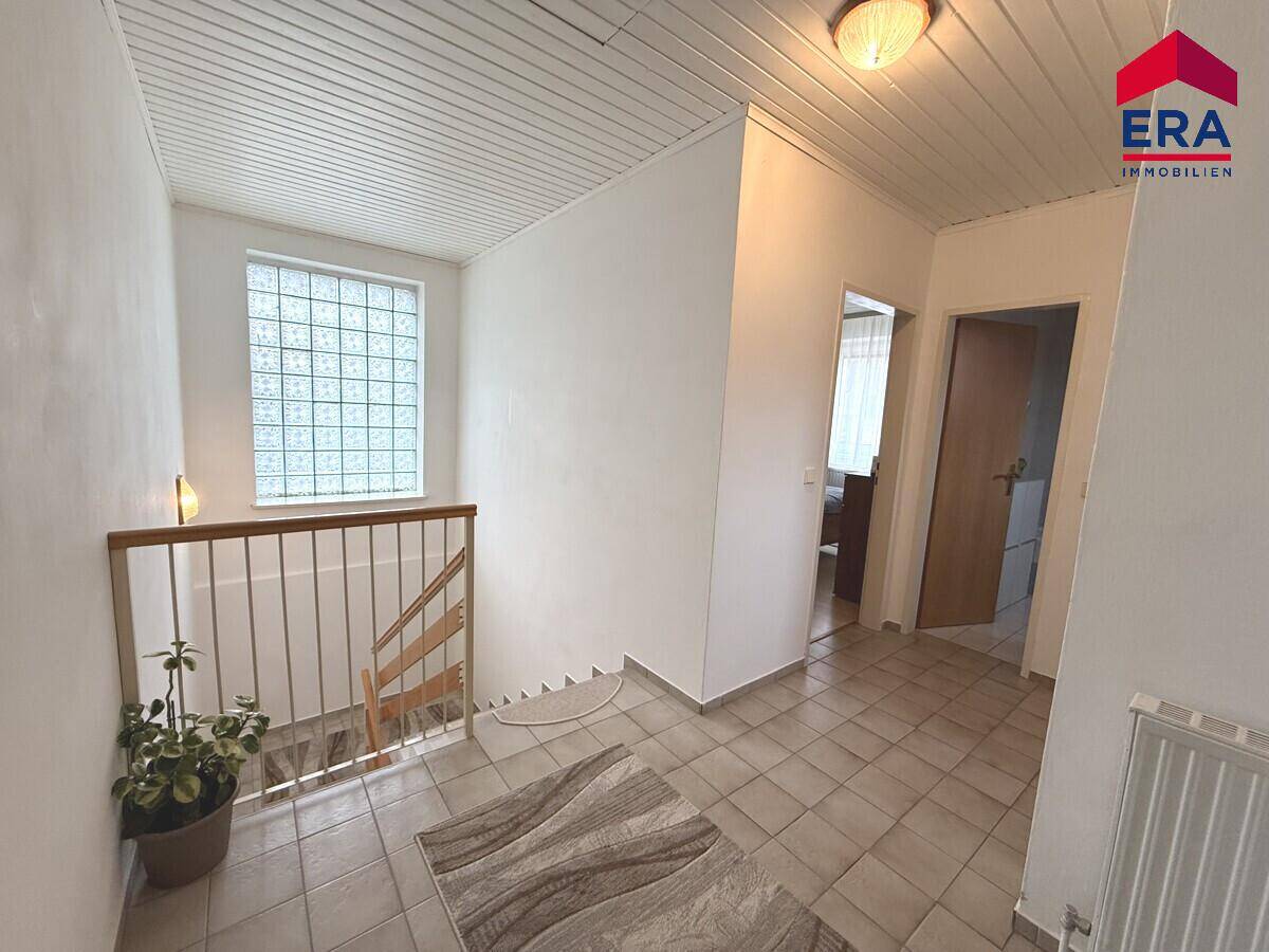 Heller Flur mit Fliesenboden, Treppenaufgang und einem Fenster aus Glasbausteinen.