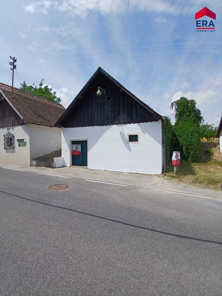 Ansicht eines charmanten alten Gebäudes mit weißer Wand und dunklem Giebeldach, umgeben von ländlicher Vegetation.