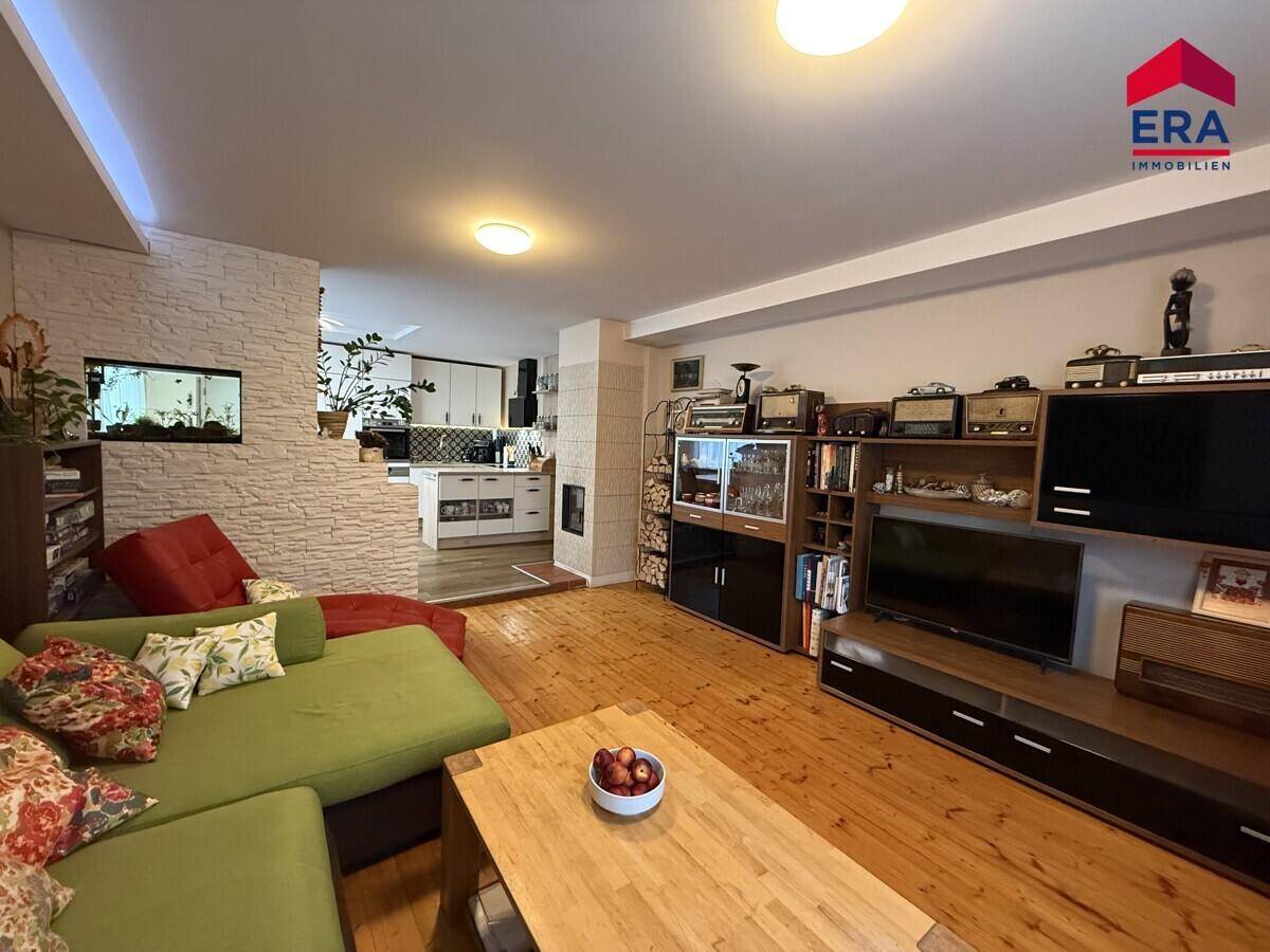 Gemütliches Wohnzimmer mit grünem Sofa, Holzboden und einem Medienbereich, offen zur Küche im Hintergrund.