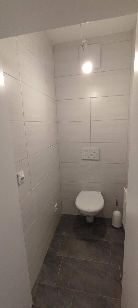 Separates WC mit wandhängender Toilette, weißen Wandfliesen und dunklem Fliesenboden.