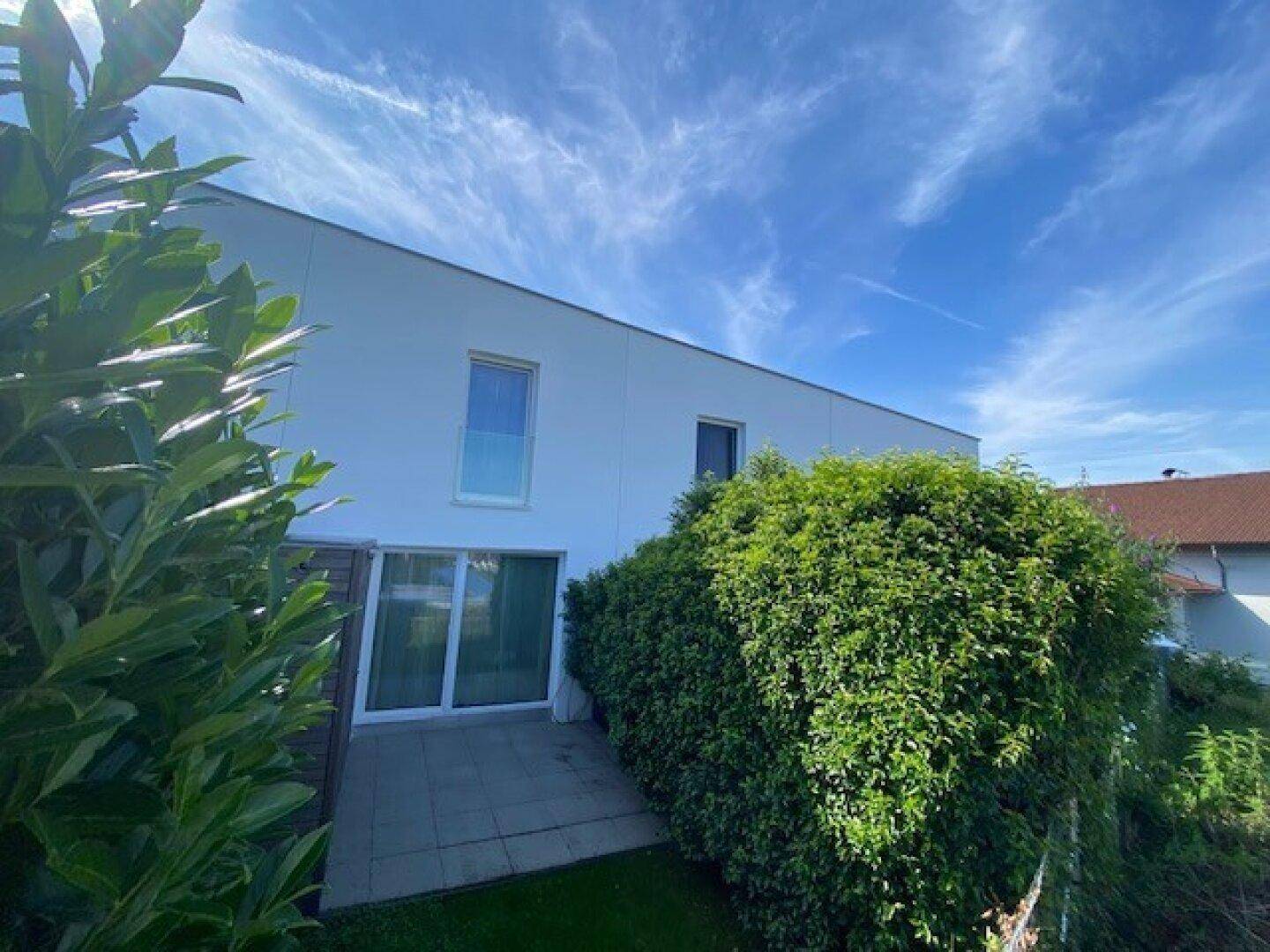 Moderne Gebäudefassade mit kleiner Terrasse, umgeben von einer dichten grünen Hecke unter blauem Himmel.
