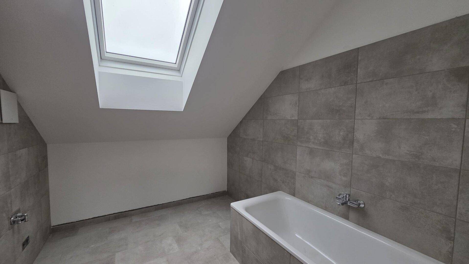 Modernes Badezimmer im Dachgeschoss mit Badewanne, grauen Fliesen und Dachfenster für natürliches Licht.