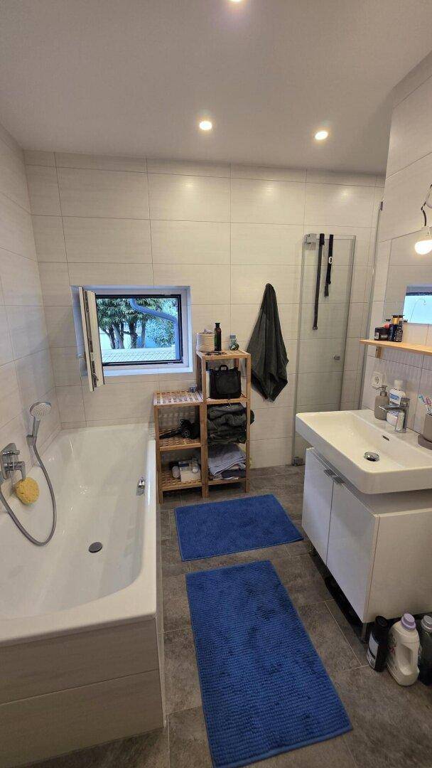 Helles Badezimmer mit Badewanne, Fenster und Blick ins Grüne, ausgestattet mit blauen Badematten.