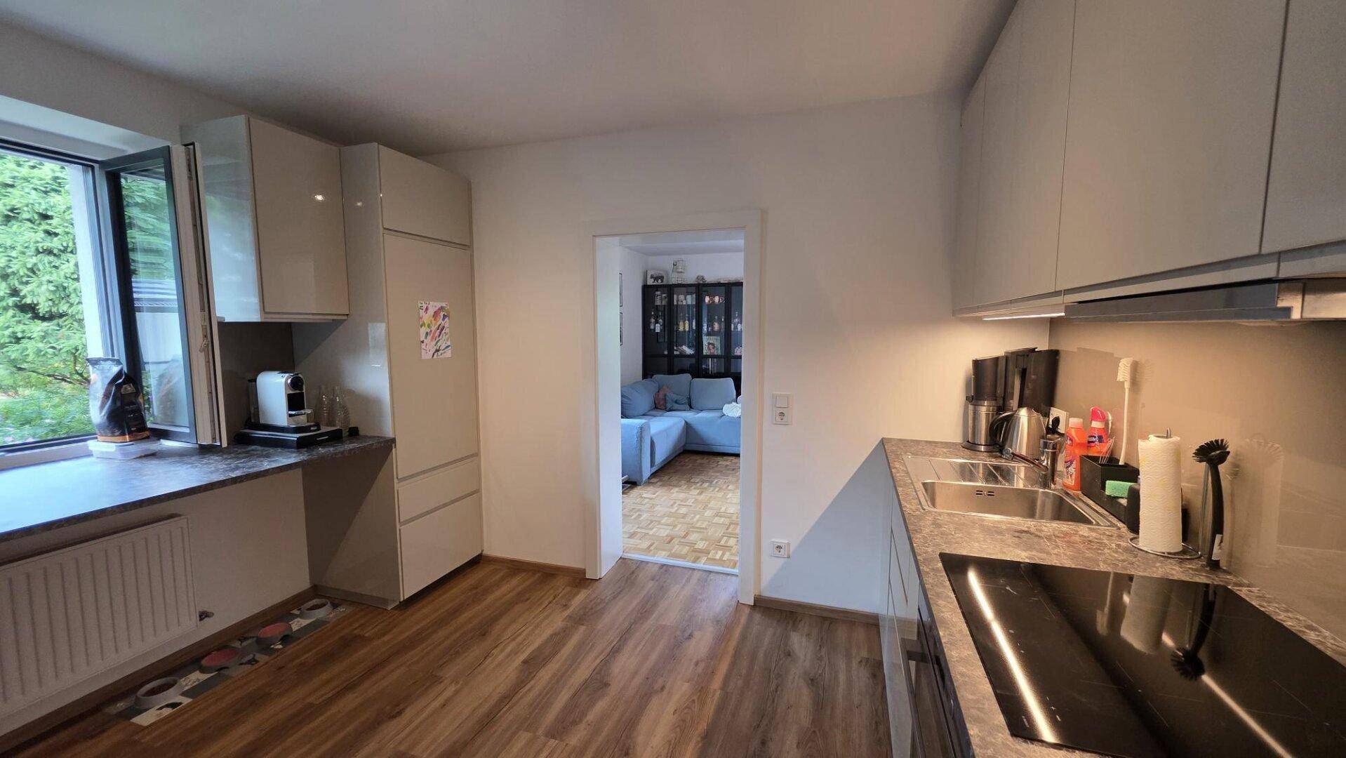 Funktionale Küche mit Fenster, Kaffeemaschine und Blick ins Grüne sowie Zugang zum Wohnzimmer.