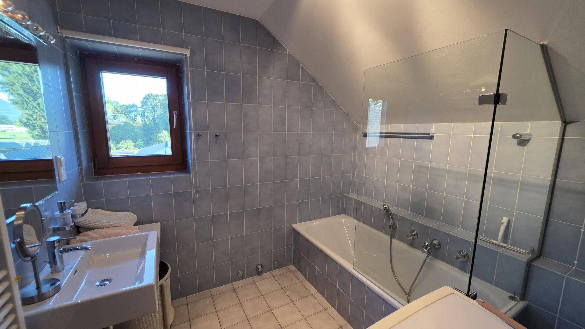 Badezimmer mit blauen Fliesen, Badewanne mit Duschabtrennung und Fenster.