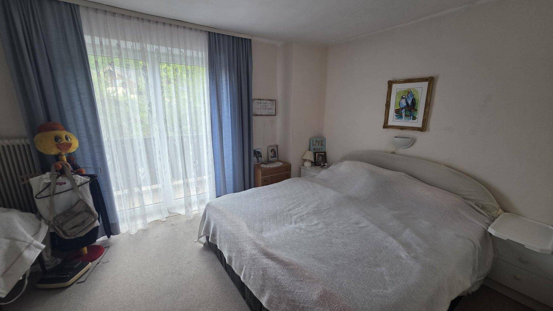 Schlafzimmer mit Doppelbett, Nachttischen und einem großen Fenster mit blauen Vorhängen, das viel Licht spendet.