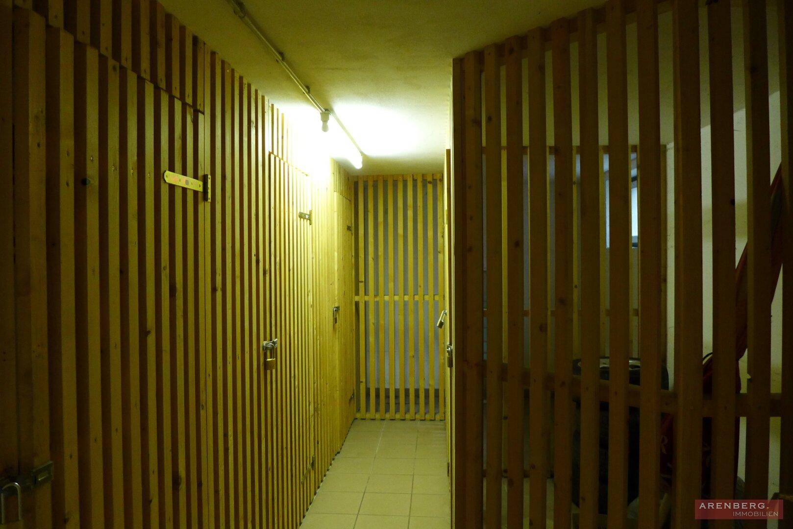 Kellerflur mit Holzverschlägen für Abstellräume, beleuchtet von Neonröhren.