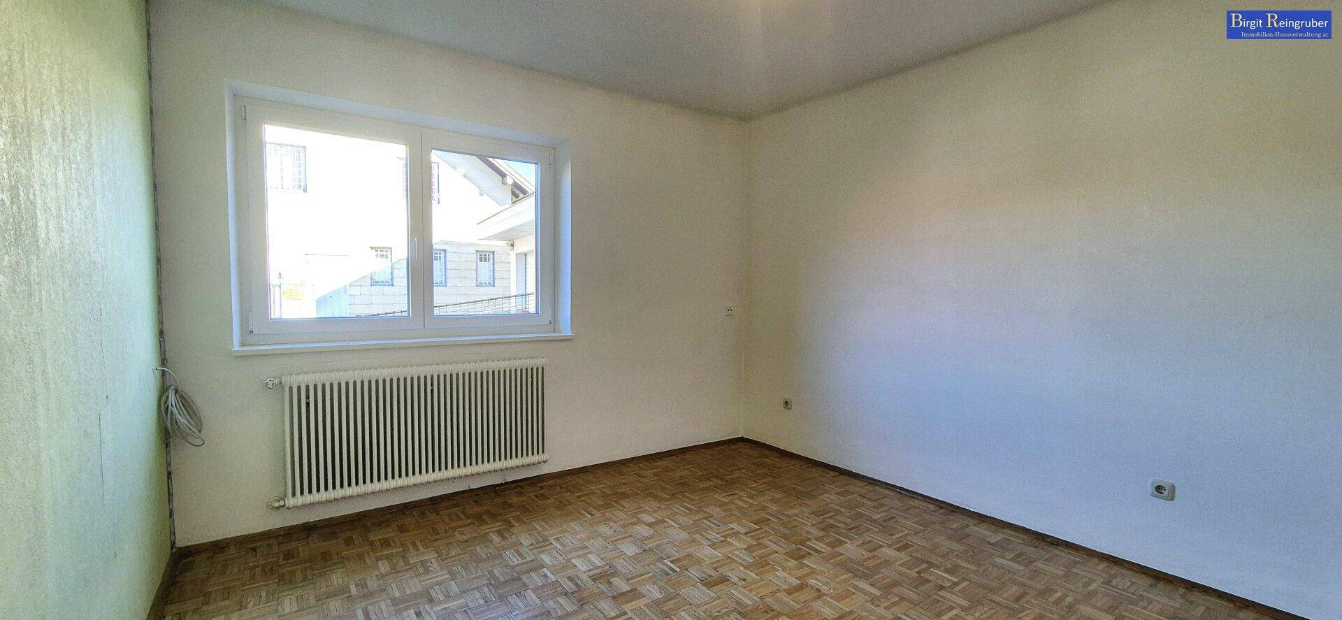 Leerer Raum mit Parkettboden, großem Fenster und Heizkörper.