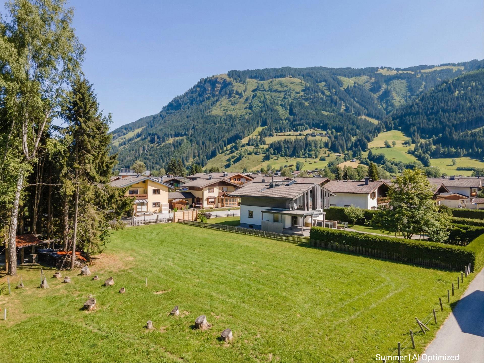 Weitläufige Grünfläche mit Wohnhäusern und bewaldeten Bergen im Hintergrund unter klarem Sommerhimmel.
