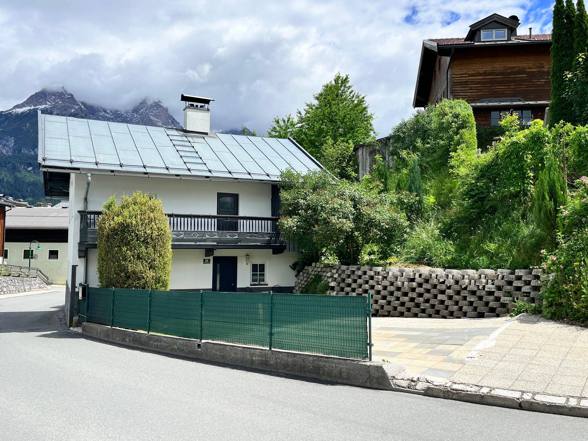 Hausfassade mit Balkon und grüner Umzäunung, im Hintergrund sind Berge sichtbar.