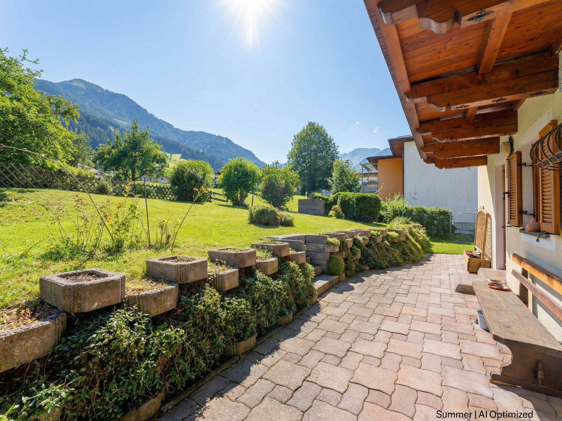 Sonniger Gartenbereich mit gepflastertem Weg, Rasenfläche und Blick auf die umliegenden Berge.