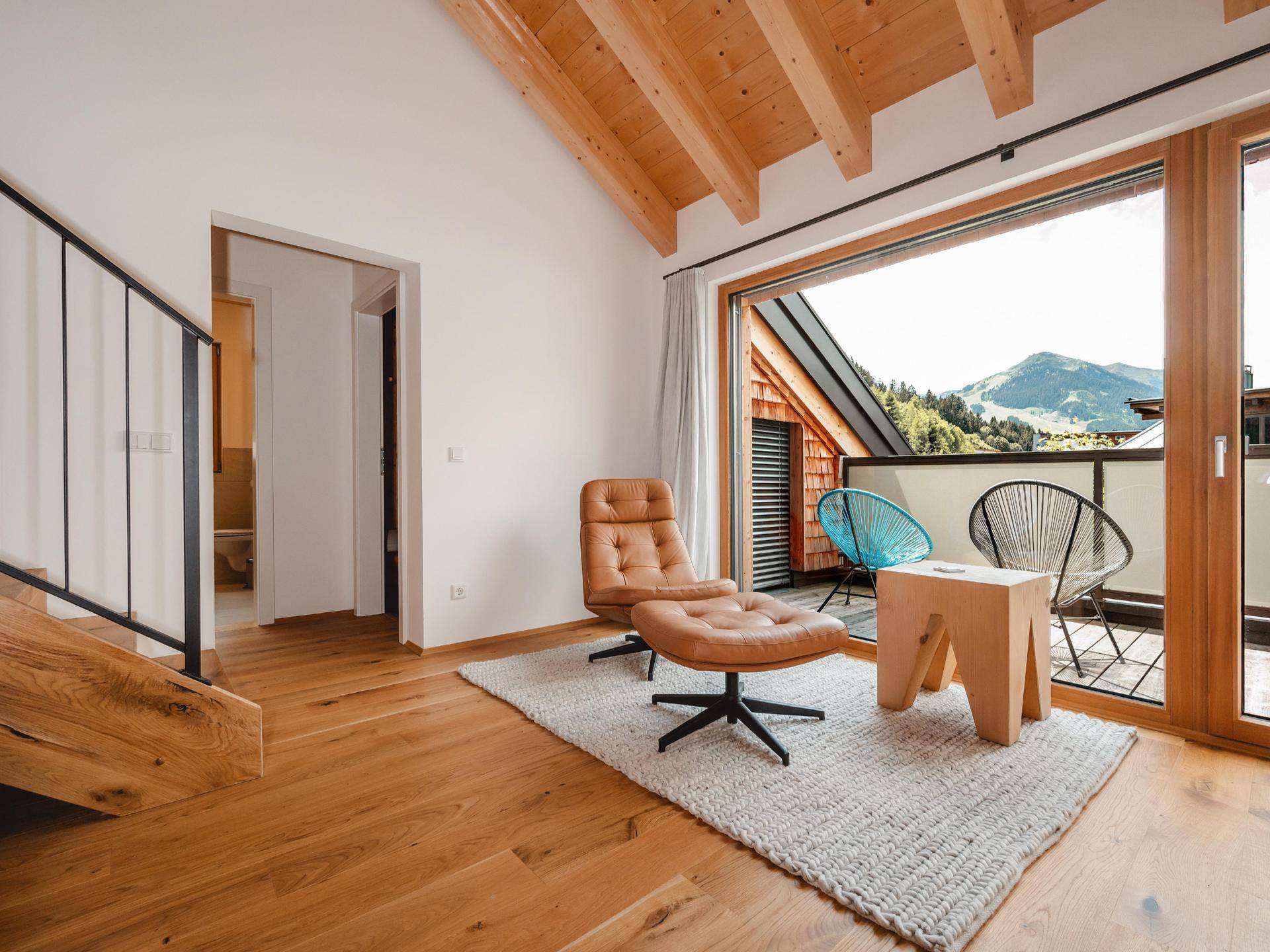 Gemütlicher Wohnbereich mit Ledersessel, Holzbalkendecke und Zugang zu einem Balkon mit Bergblick.
