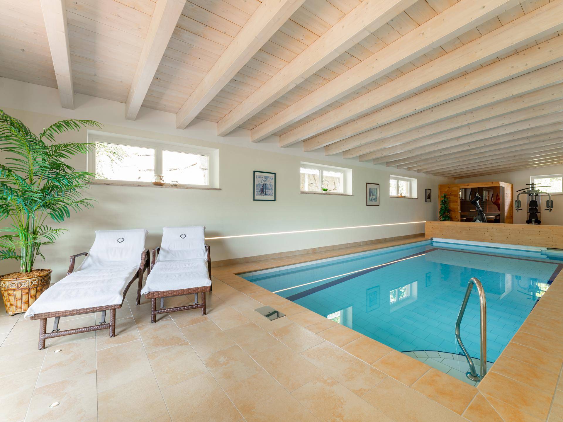 Heller Innenpoolbereich mit Holzbalkendecke, Liegestühlen und Fitnessgeräten im Hintergrund.